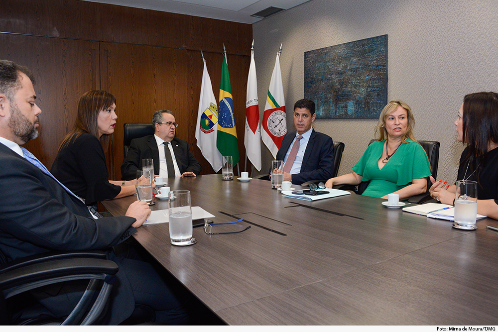 Autoridades em mesa retangular com bandeiras de Minas e do Brasil ao fundo