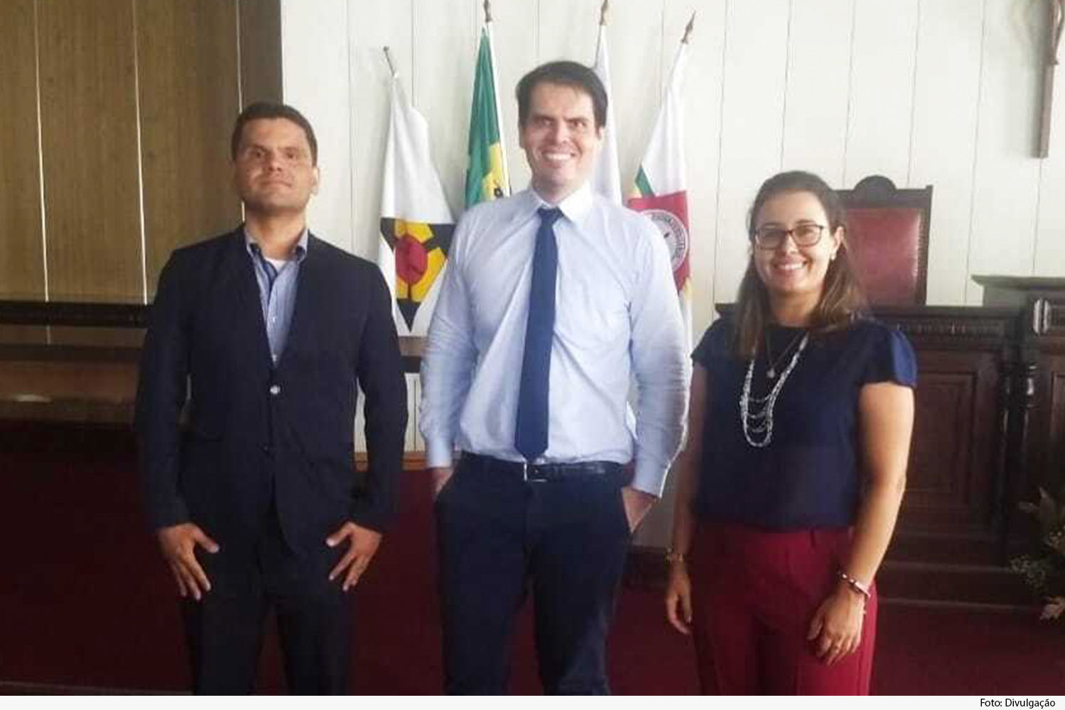 Três pessoas de pé, tendo bandeiras de Minas, do Judiciário e do município ao fundo