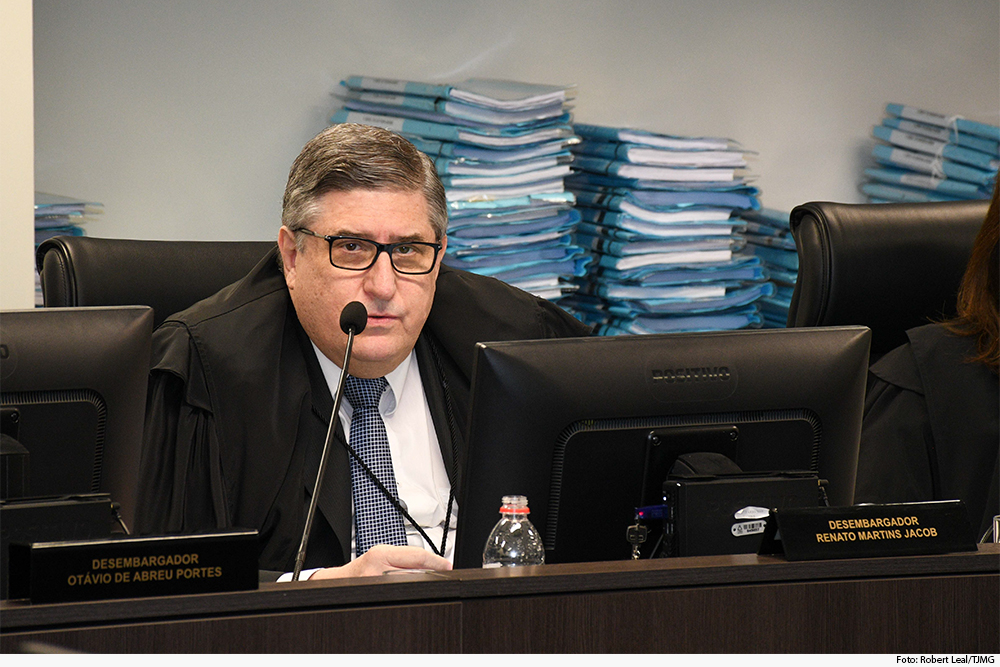 Homem fala ao microfone em sessão de julgamento, com processos ao fundo