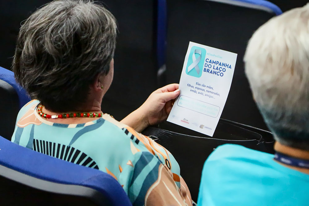 Mulher idosa, sentada na plateia, observa folder sobre a campanha