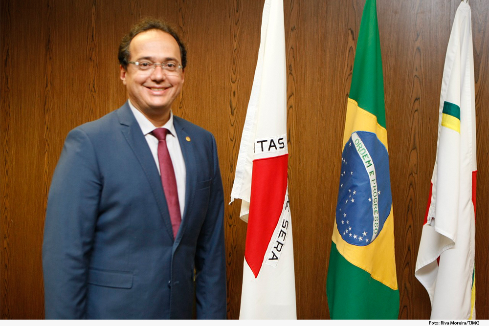 Magistrado de pé tendo bandeiras do Brasil, Minas e TJMG