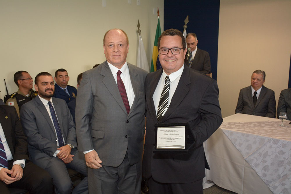 Servidor ao lado de juiz mostra placa de homenagem em solenidade