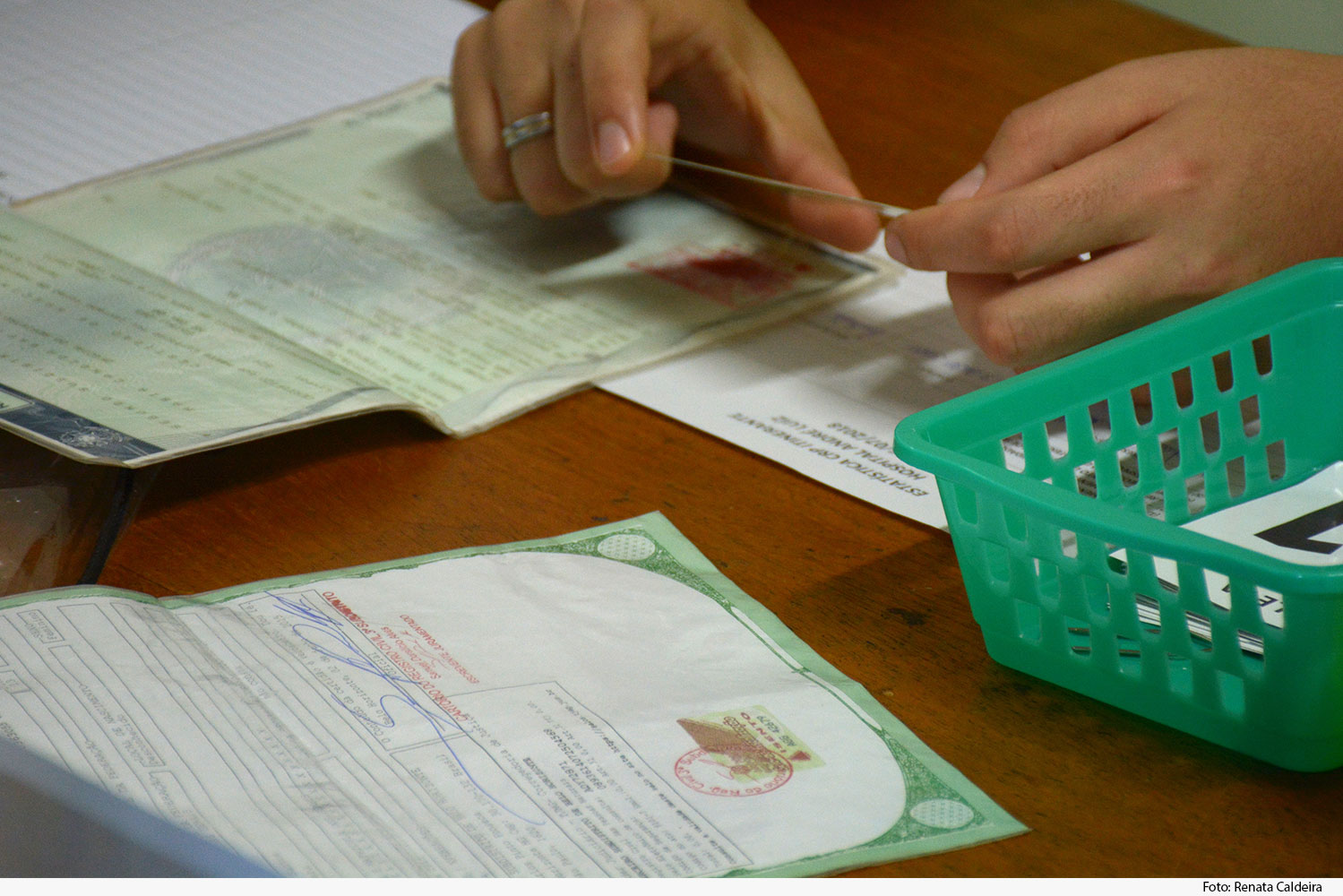 Mão segurando carteira de identidade em cima de mesa com vários documentos e cesto de plástico verde