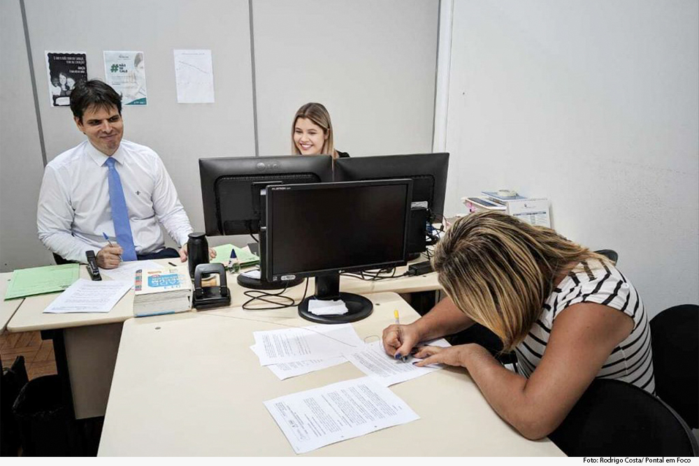 Juiz, assentado em mesa, ao lado de servidora, observa mulher assinando documento