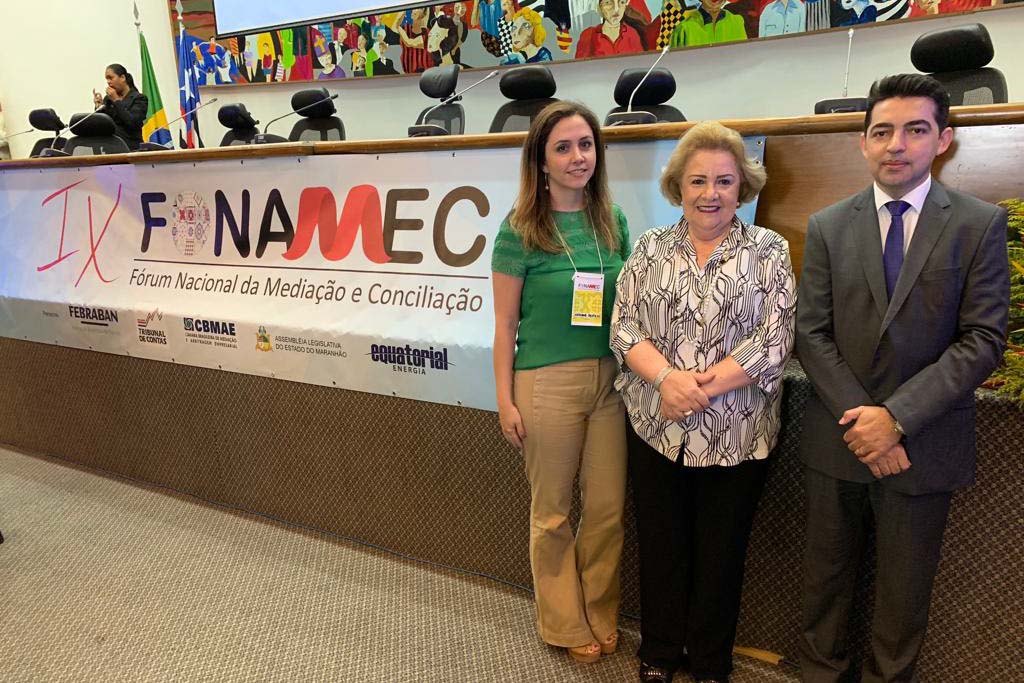 Homem e duas mulheres posam em frente a banner de evento em auditório