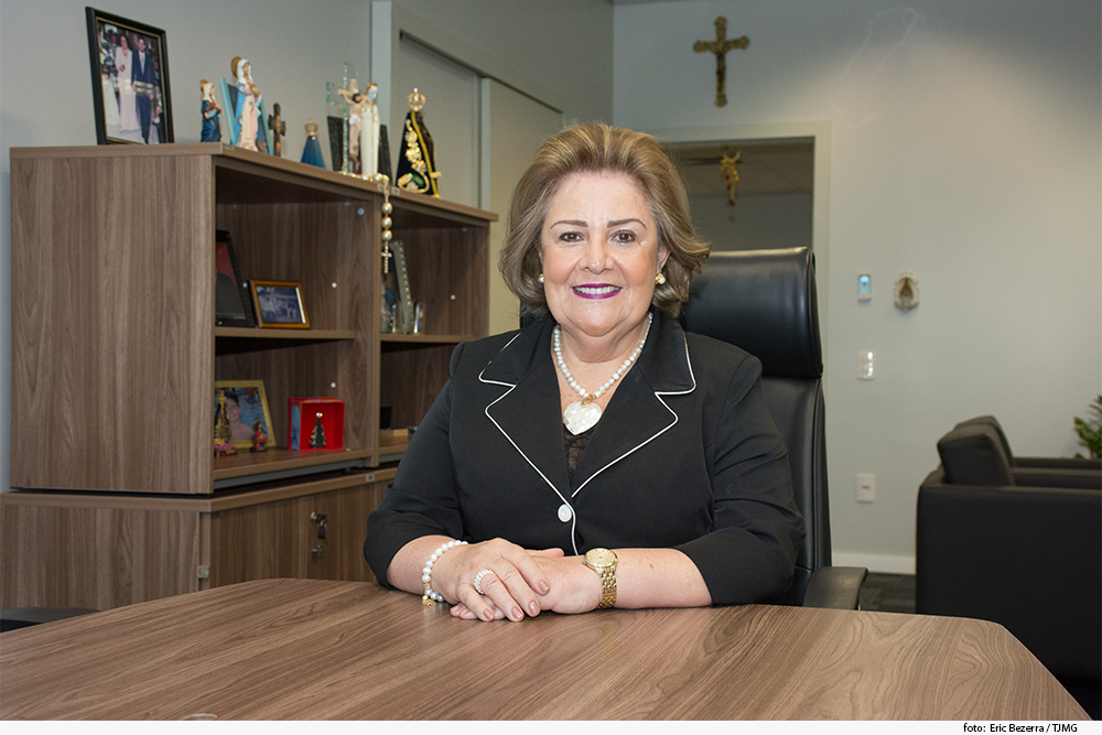 Magistrada assentada com as mãos cruzadas sobre a mesa, em gabinete com livros ao fundo