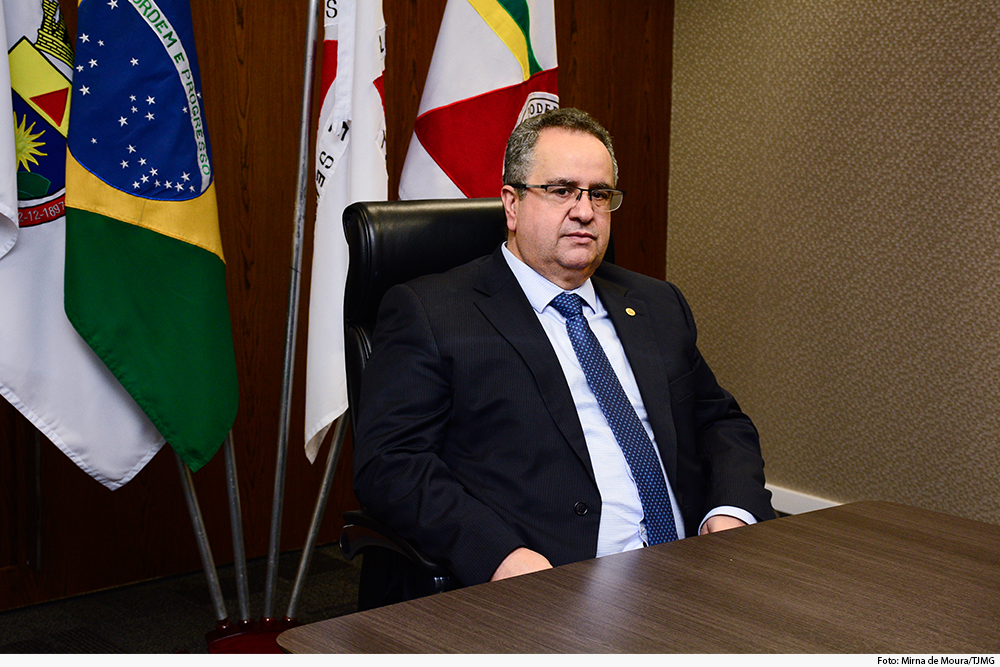 Homem de terno sentado à mesa tendo bandeiras de Minas, do Judiciário e do Brasil ao fundo