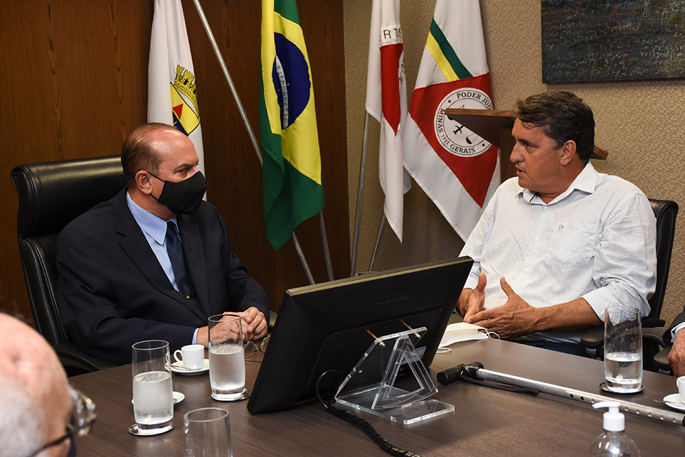 Magistrado em mesa de reunião com autoridades, tendo bandeiras ao fundo