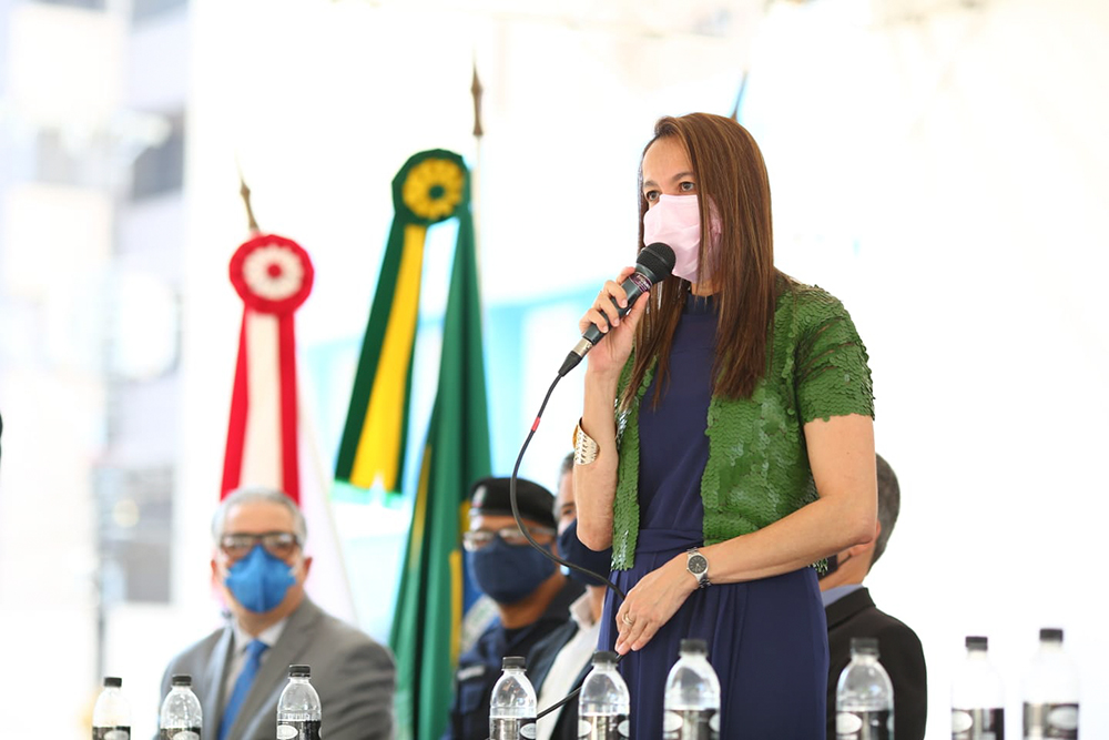Juíza, de pé, se pronuncia em solenidade, tendo autoridades e bandeiras ao fundo