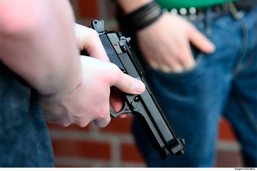 Imagem mostra as pernas e mãos de dois homens, um dos quais manipula uma pistola