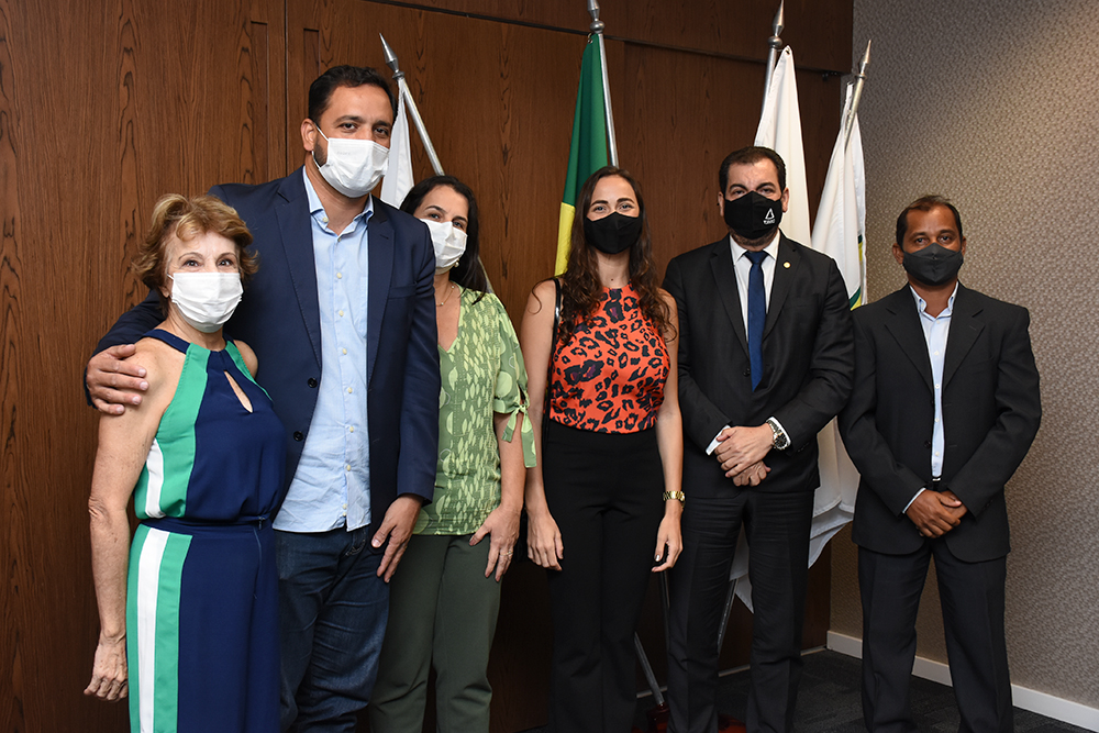 Grupo de autordades, de pé, em sala de reunião, com bandeiras ao fundo