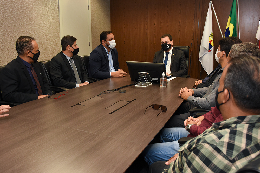 Ao lado do presidente, deputado, em mesa de reunião, fala aos presentes, 